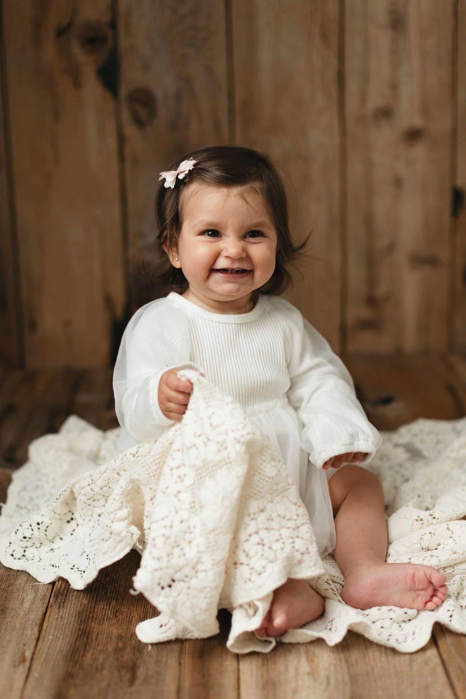 Smiling baby girl in white knit dress sitting on lace blanket against rustic wood background. Elizabeth Lang Photography Surrey