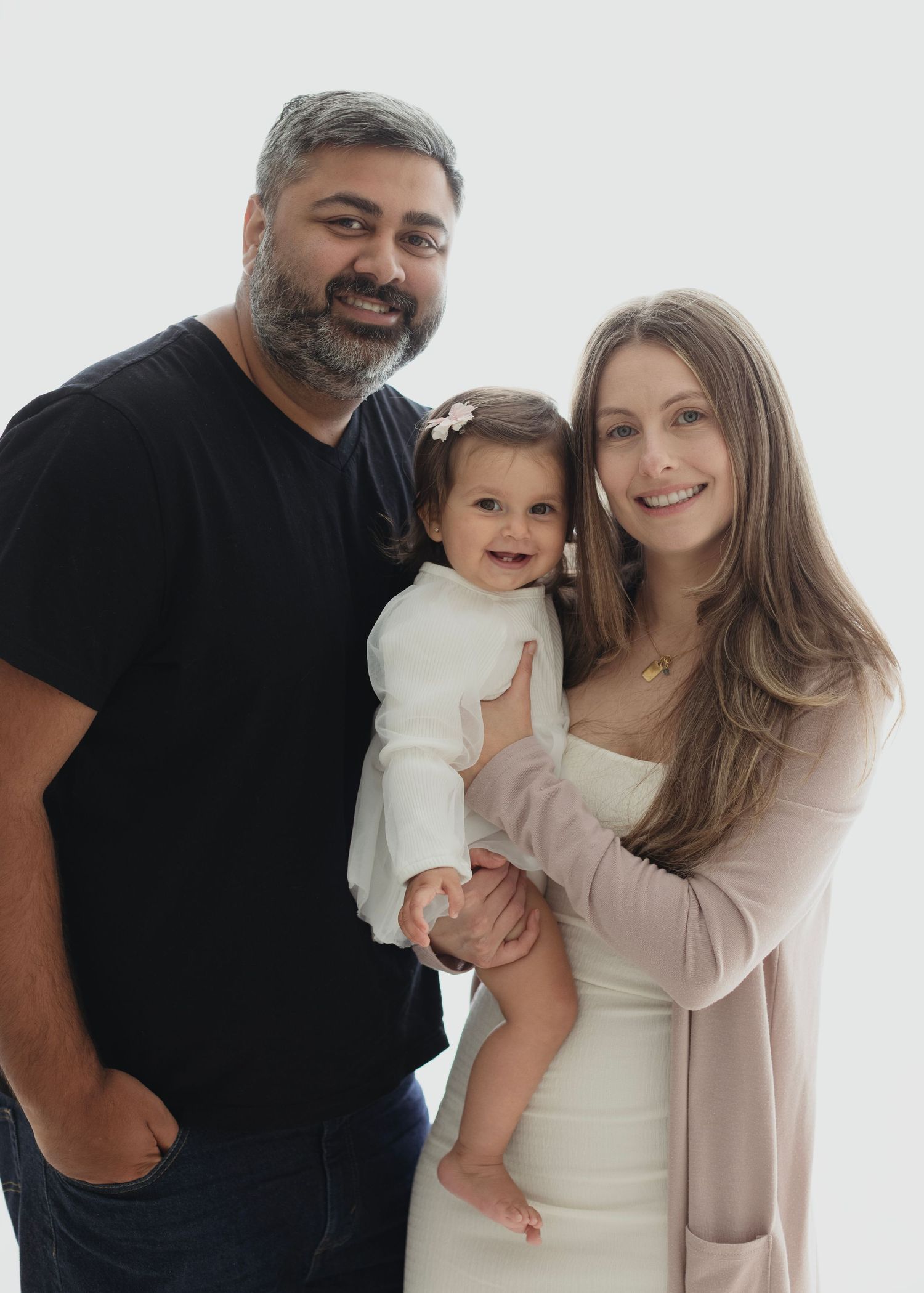 A smiling family of three poses together, with parents holding their happy baby girl dressed in white against a bright background. Elizabeth