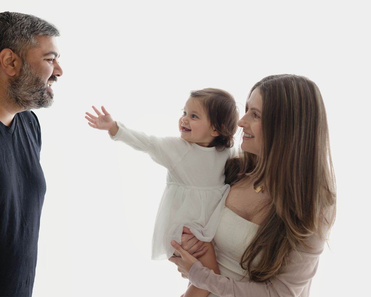A happy family with a bearded father, smiling mother, and a baby girl in a white dress reaching out joyfully. Elizabeth Lang Photography Sur