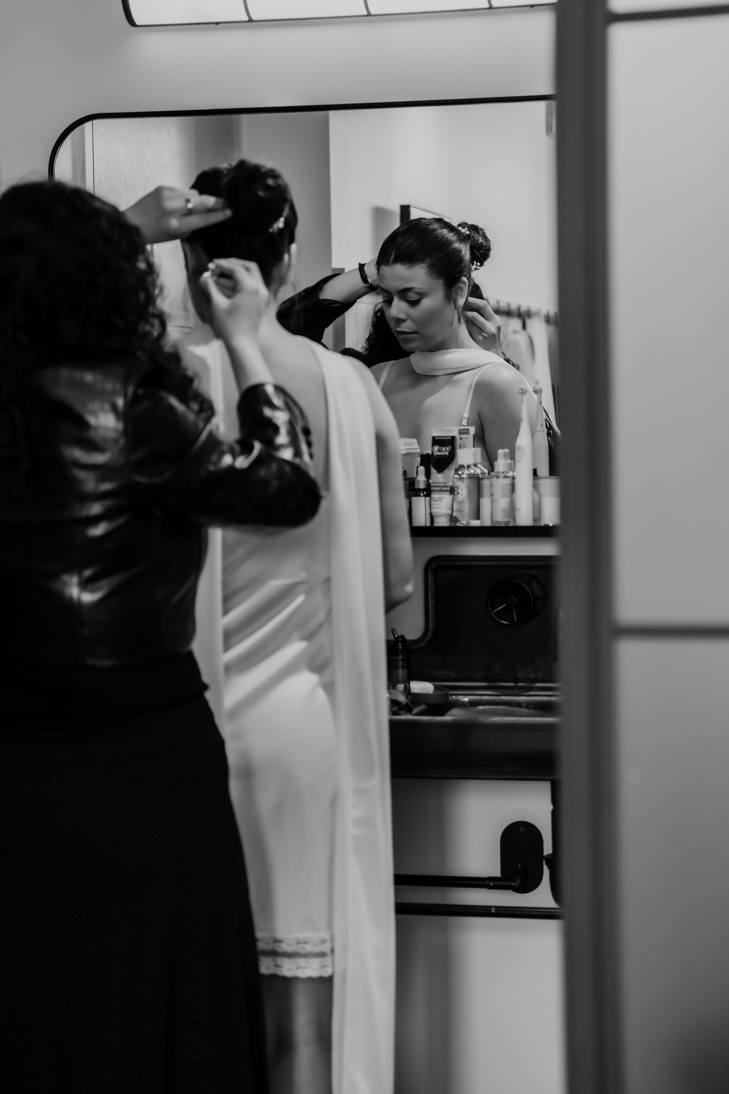 Bride getting hair styled by assistant in mirror reflection, black and white wedding preparation photo.