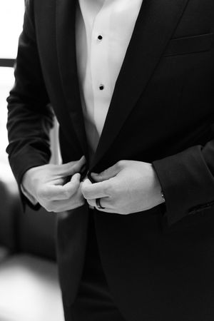 Close-up black and white photo of groom buttoning his black tuxedo jacket on wedding day.