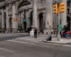 Wedding couple in formal attire crosses busy New York City street in front of grand arched building.