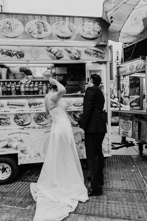 Bride and groom in wedding attire ordering food from NYC street cart, black and white photo.