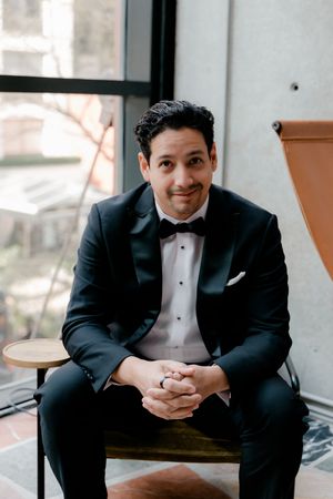 A groom in a black tuxedo with bow tie sits on a wooden stool, smiling with hands clasped near a large window.