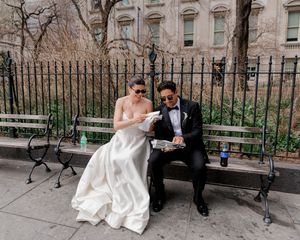 Bride and groom in wedding attire eating food on a park bench in New York City.