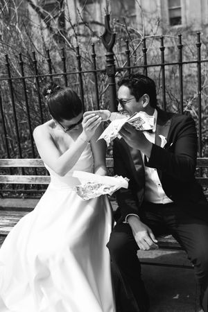 Black and white photo of newlyweds sharing pizza on a park bench in wedding attire.