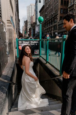 Bride and groom holding hands at NYC 28th Street subway station entrance, bride in white gown looking back smiling.