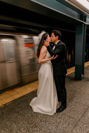 Newlyweds share romantic kiss on NYC subway platform as train blurs past in motion behind them.