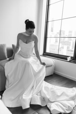 Black and white photo of bride in strapless satin gown sitting by window adjusting her dress.