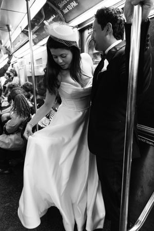 Black and white photo of bride and groom riding NYC subway, bride in white gown and veil hat, groom in black suit.