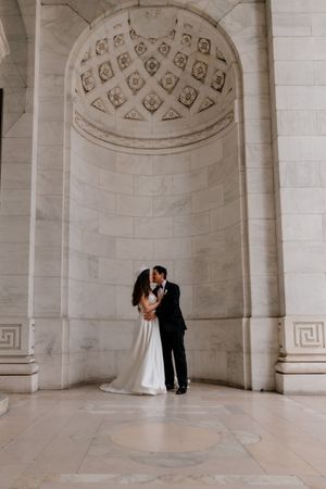 Bride and groom kissing beneath a grand marble arch in an elegant neoclassical building.