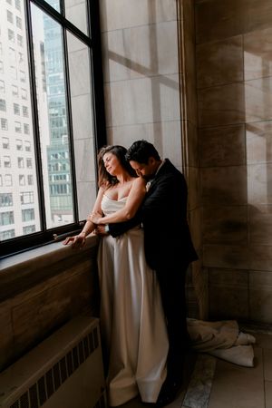 Bride and groom embracing by tall window in elegant stone building, city skyline visible, intimate wedding portrait.