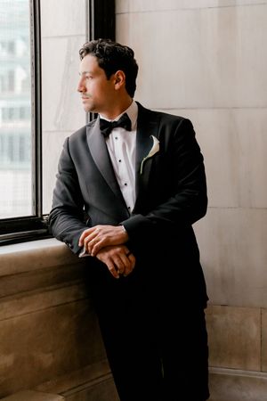 Dapper groom in black tuxedo with boutonniere gazing thoughtfully out window on wedding day.