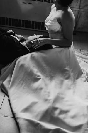 Black and white photo of bride in strapless gown sitting beside groom in tuxedo on floor, holding hands.