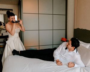 Bride in white gown photographing groom lying on hotel bed in white shirt during wedding day celebration.