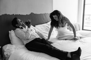 Black and white photo of a couple in wedding attire relaxing on a hotel bed, sharing a joyful moment.