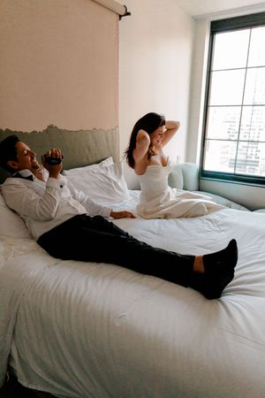 Couple in wedding attire relaxing on a white hotel bed near a city view window, groom holding a drink.