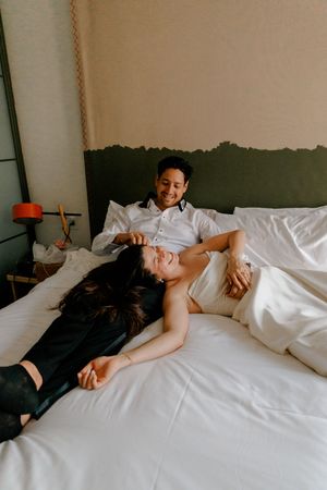 Happy newlywed couple laughing and relaxing together on white hotel bed after wedding day.