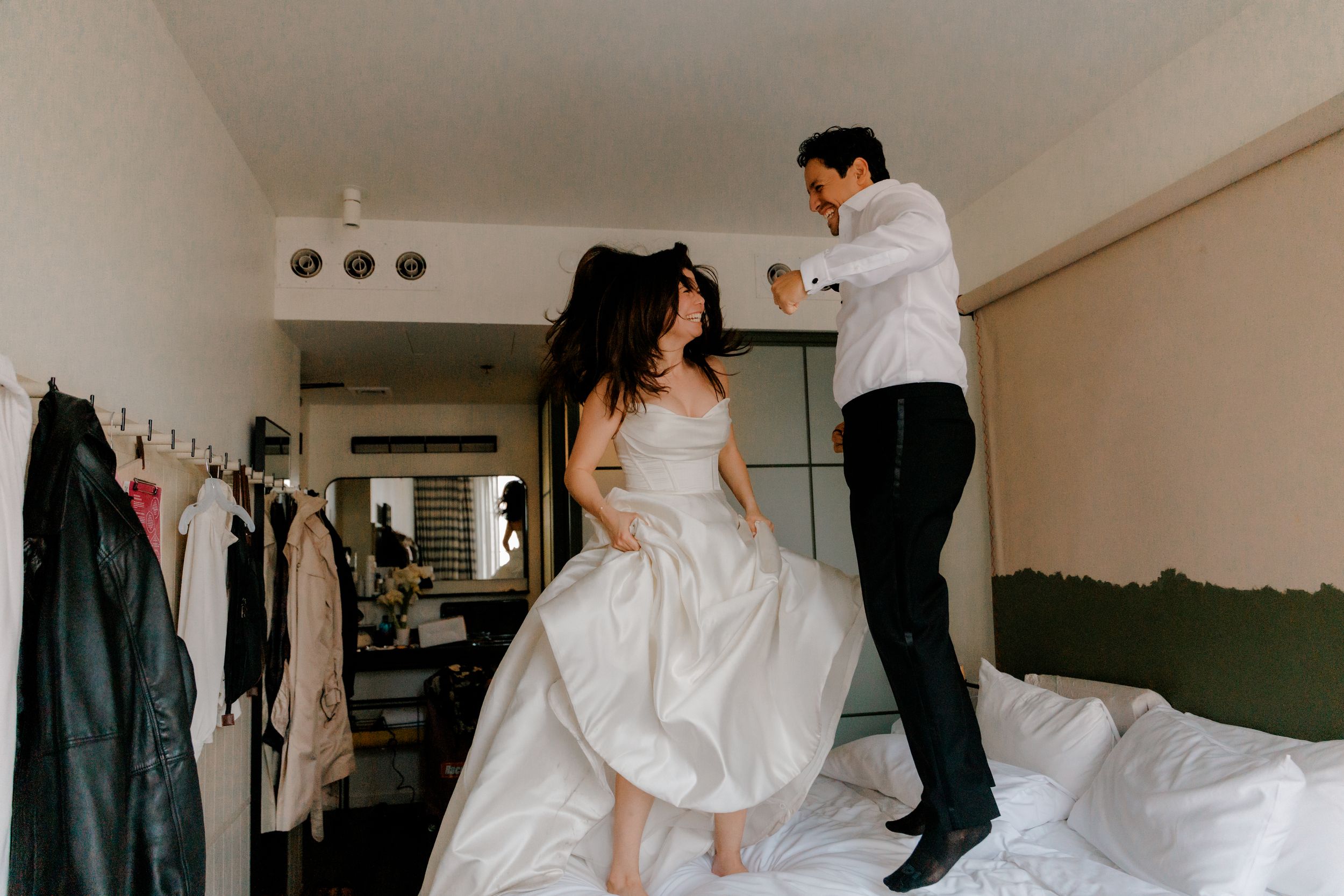 Joyful newlyweds jump and dance on hotel bed in wedding attire celebrating their special day.