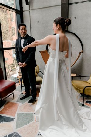 Bride in elegant white gown extends hand to groom in black tuxedo during wedding first look in modern venue.
