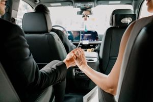 Couple holding hands in luxury black car backseat, bride in white dress and groom in suit, wedding day transportation.