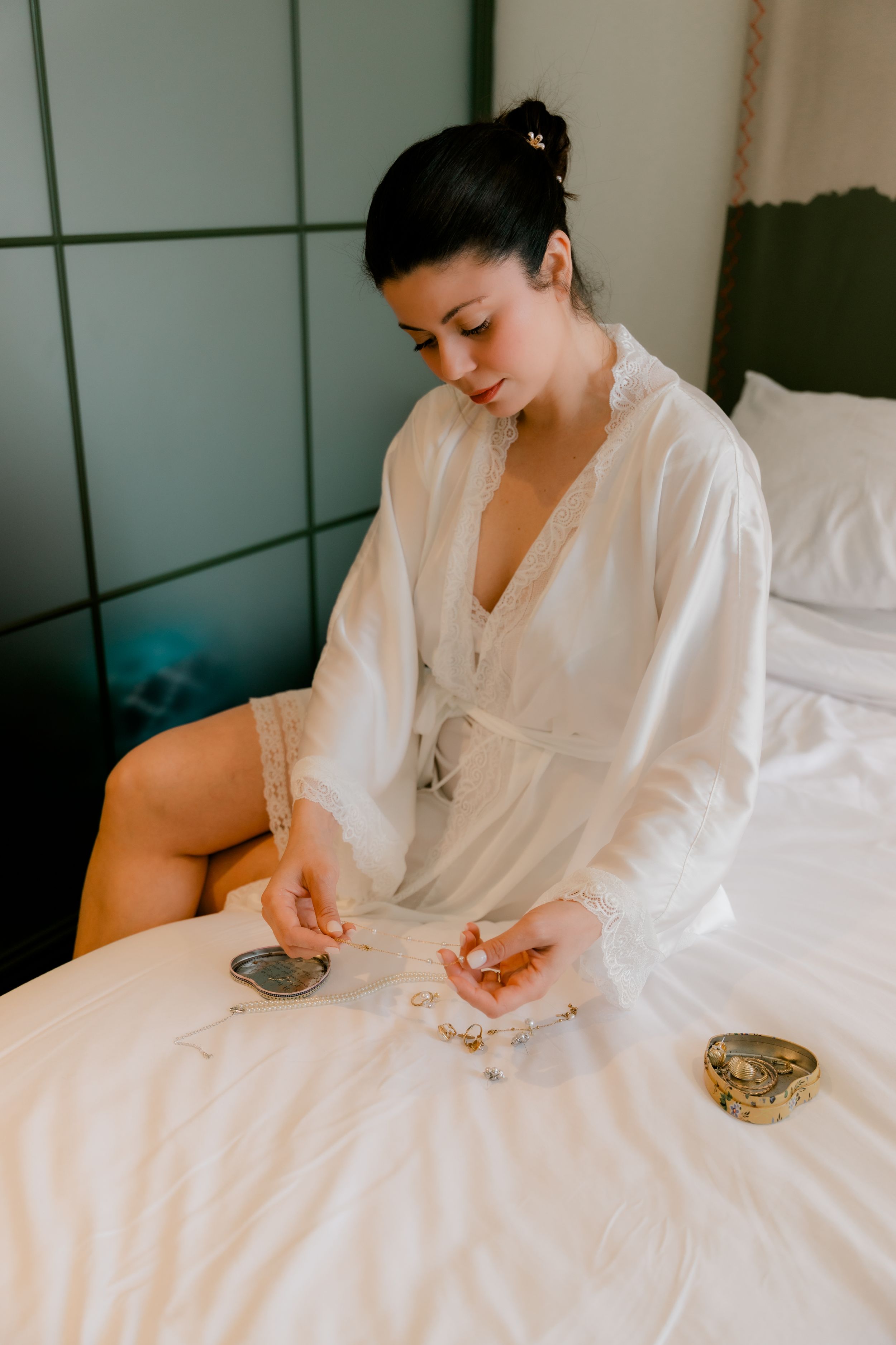 Bride in white lace robe sitting on bed examining jewelry before wedding ceremony.