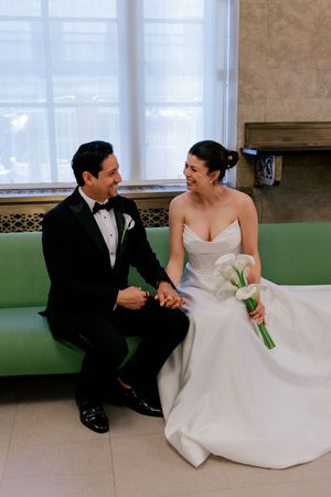Bride and groom sitting on green bench, holding hands and laughing together after their ceremony.