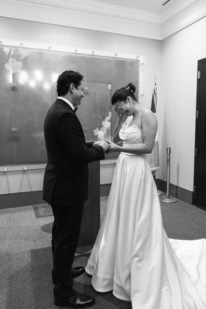 Emotional bride wipes tears as groom places ring on her finger during intimate black and white wedding ceremony.
