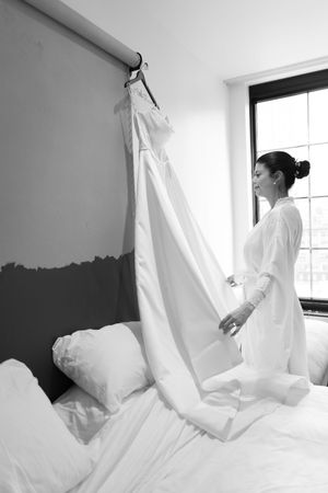 Bride in white robe kneeling on bed touching her wedding dress hung on wall.