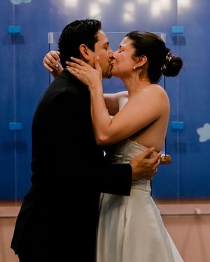 Bride and groom share a romantic first kiss in wedding attire, with a blue decorative backdrop behind them.