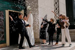 Newlywed couple exits building as guests toss white confetti in joyful outdoor wedding celebration.