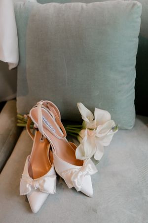 White bridal heels with bow and pearl detail beside white calla lilies on a velvet chair with mint cushion.