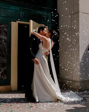 Newlywed couple sharing a romantic kiss outside a church entrance with colorful confetti falling around them.