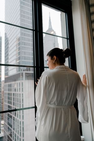 Bride in white silk robe gazing out hotel window at NYC skyline with Empire State Building view.
