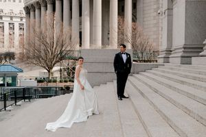 Bride in white gown and groom in tuxedo pose on steps of grand neoclassical building for wedding portrait.