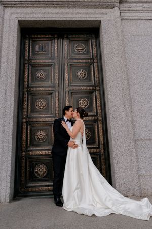 Bride and groom sharing a kiss in front of ornate bronze doors at a grand stone building.