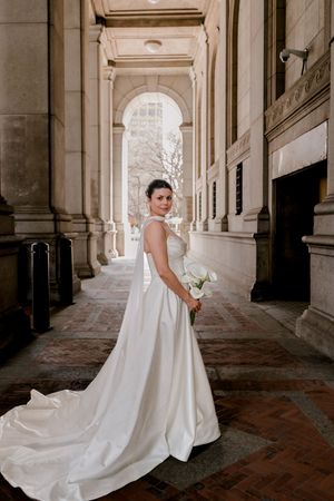 Bride in stunning white satin gown with long train poses among grand stone columns holding white bouquet.