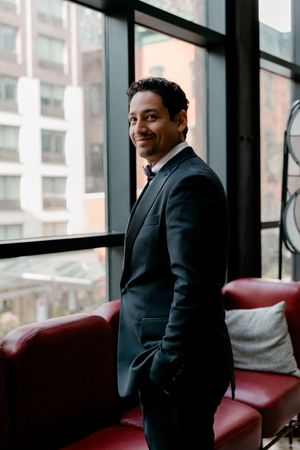 Smiling groom in black tuxedo standing by large windows with city views in a modern venue.