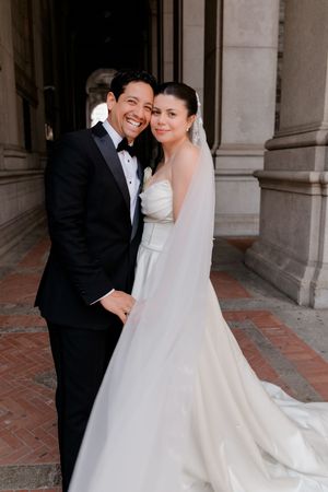 A smiling bride and groom pose together in elegant wedding attire outside a grand stone building.