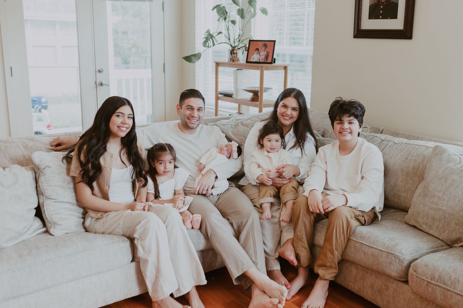 Happy family of six posing together on a couch in a bright living room with a newborn baby.