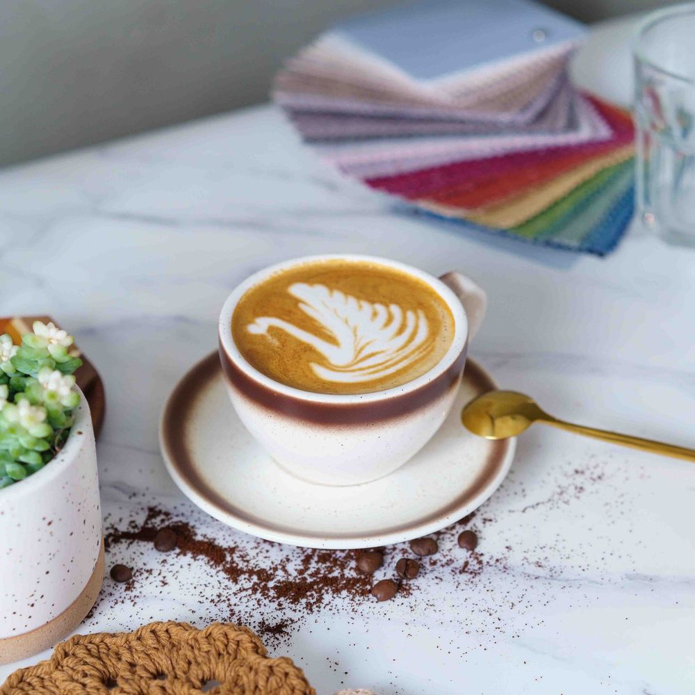 A cup of coffee with latte art sits on a saucer next to scattered coffee grounds and colorful paper swatches.