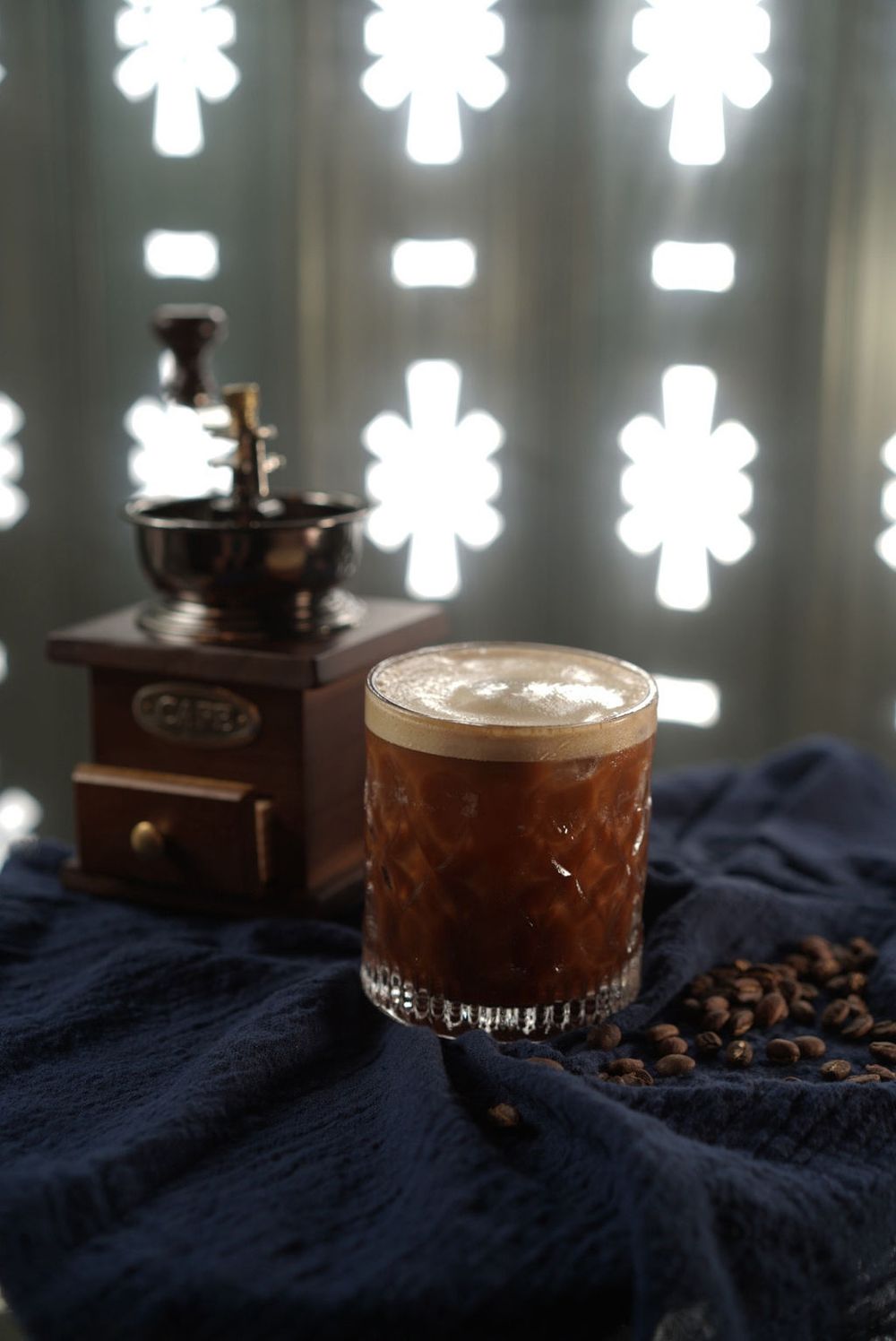 Coffee beans and vintage grinder with creamy espresso drink in glass against dark blue fabric and glowing lights.