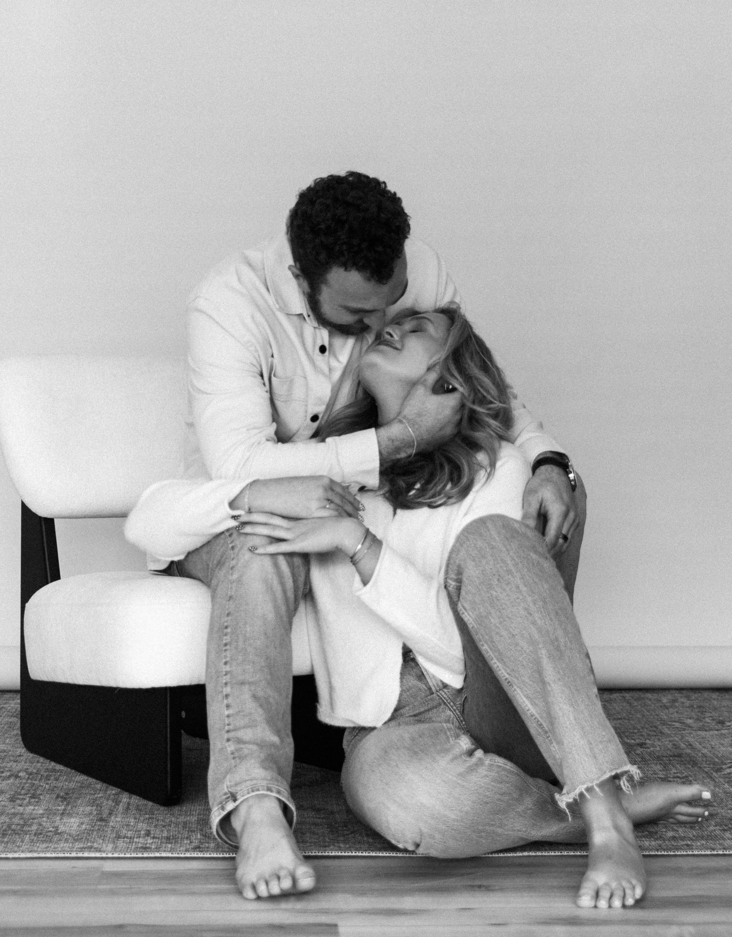 Black and white photo of a couple sharing a tender embrace on the floor, leaning against a modern white chair.