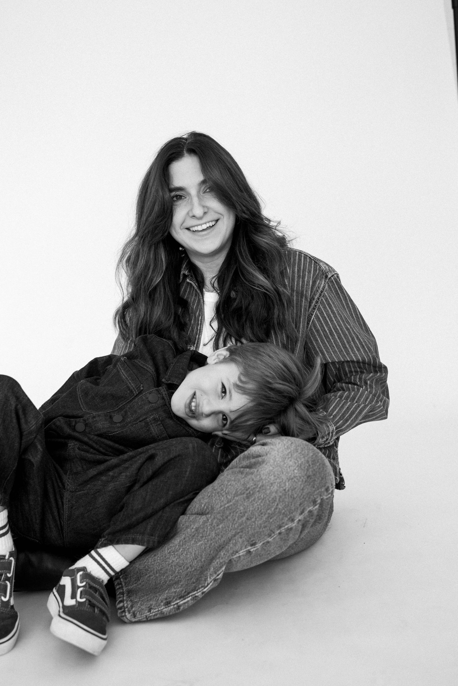 Black and white photo of a smiling woman with long hair sitting on floor with a young child leaning on her.