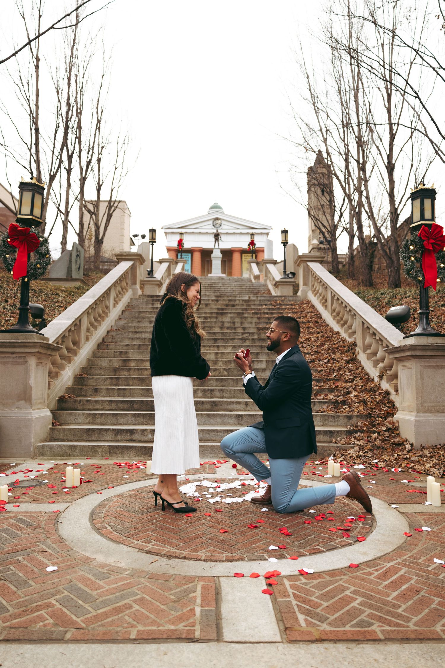 A couple stands smiling under a stone archway entrance, man in navy blazer and jeans, woman in white skirt and black fur jacket.