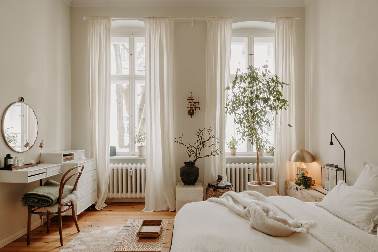 Bright Scandinavian bedroom with white curtains, wooden desk, round mirror, green plants, and cozy bedding near tall windows.