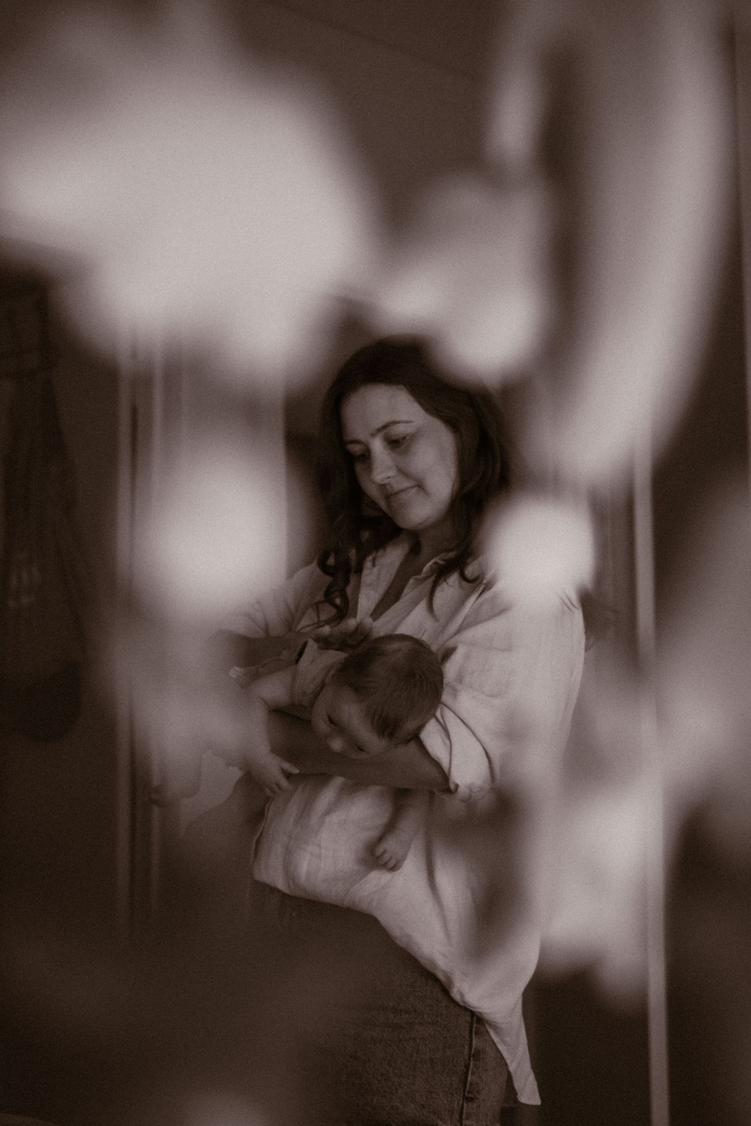 Sepia-toned photo of a mother tenderly cradling her newborn baby, shot through soft bokeh foreground elements.