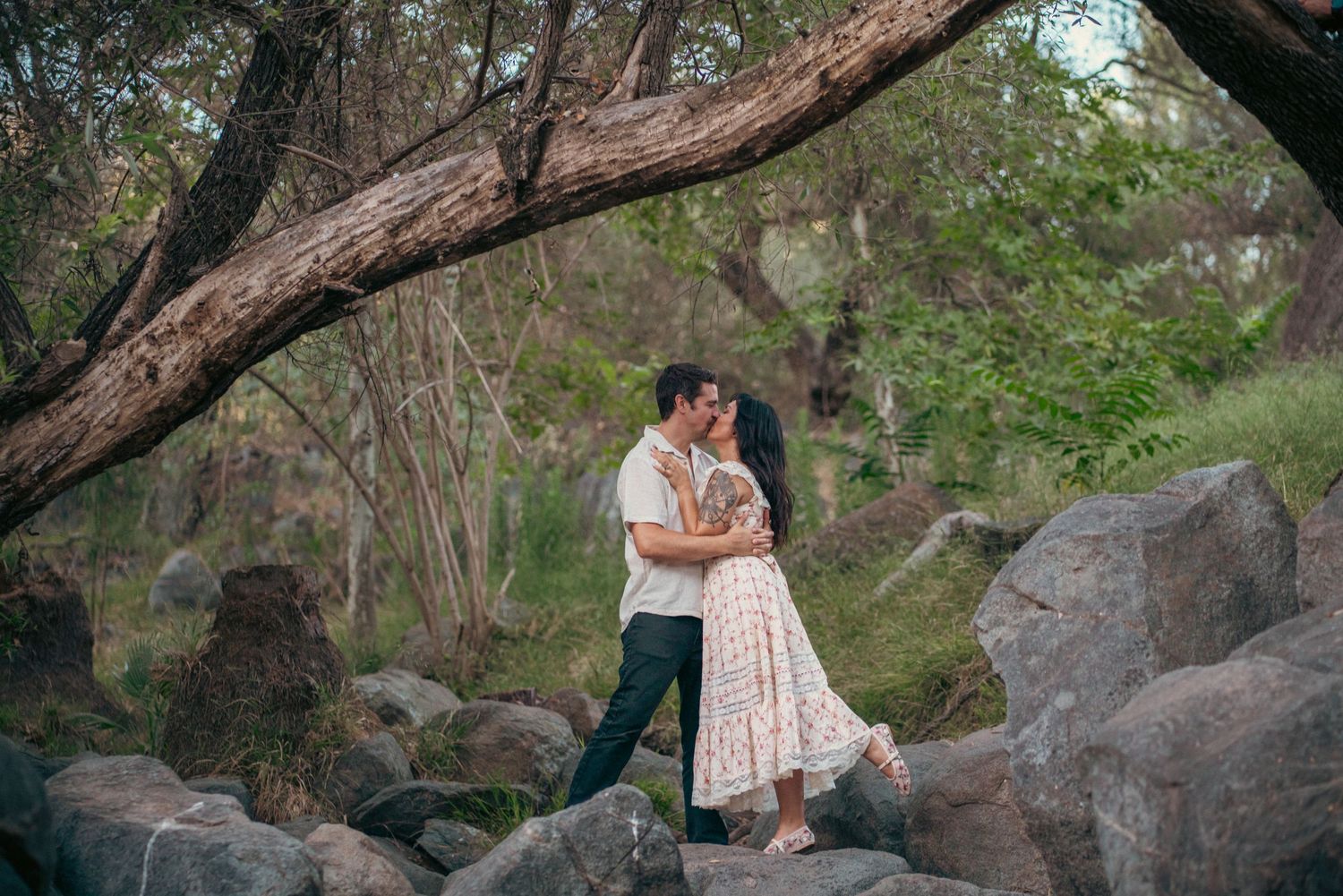 Couple embracing romantically on rocks in a lush forest setting with overhanging tree branches. Vista, CA