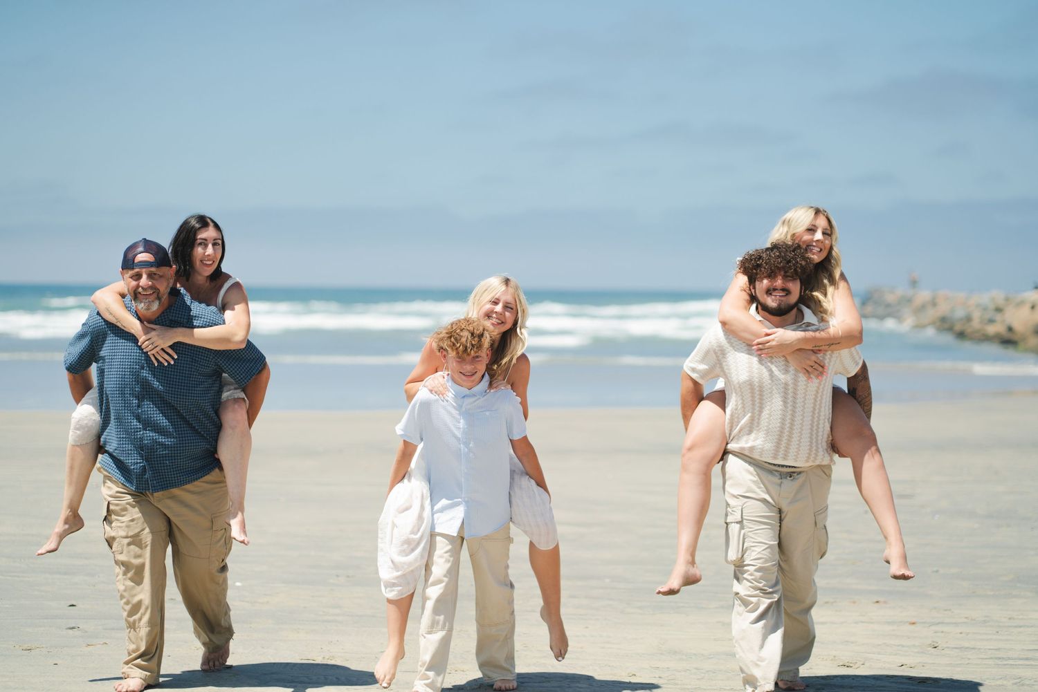 Family enjoying a sunny beach day with piggyback rides, walking on sandy shore with blue ocean waves in the background. Oceanside, CA
