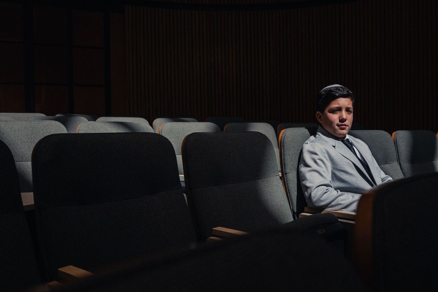 Person sitting alone in dark theater auditorium wearing gray blazer, surrounded by empty blue seats in dramatic lighting.